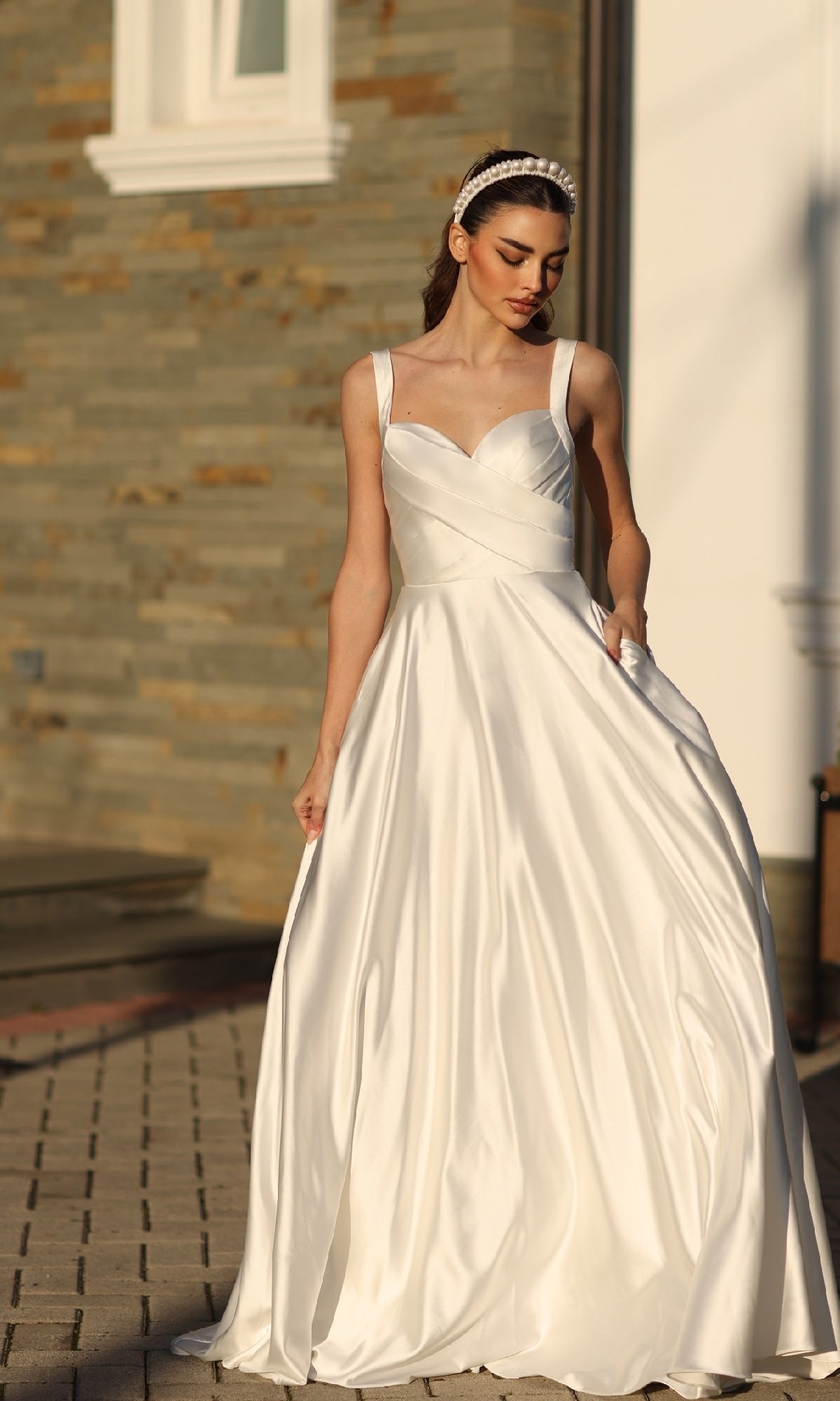 A woman wearing the Off-White Satin Prom Ball Gown: Tina Holly BB210W and a headband stands outdoors on a paved area near a stone wall.